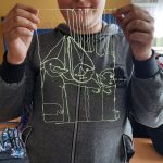 Uczeń w szarej bluzie trzyma w ręku kształt, wykonany techniką długopisu 3d, który nawiązuje do obrazu Makowskiego „Troje dzieci z chorągiewkami”.