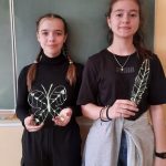 Dwie dziewczynki – obie ubrane w czarne podkoszulki, stoją na tle zielonej tablicy i trzymają w ręku wykonane przez siebie prace długopisem 3d, motyla i pióro.