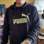 Chłopiec w bluzie dresowej z napisem ”Puma” stoi na tle klasy i trzyma w ręku wykonaną przez siebie pracę długopisem 3d.