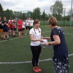 Pani dyrektor w granatowej sukience gratuluje dziewczynie dziewczynie w białej koszulce i czarnych spodniach, w tle dzieci ustawieni w czterech rzędach.