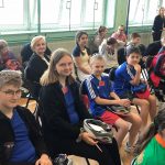 Grupa osób siedząca na krzesłach na sali gimnastycznej.