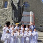 Grupa dzieci w strojach komunijnych oraz ich katecheta przed pomnikiem Jana Pawła II w Limanowej