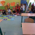 Na końcu klasy, przy kolorowej macie siedzą na podłodze trzy dziewczynki i chłopiec, obok nich na krzesłach siedzą dwie kobiety.