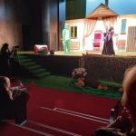 Na scenie kobieta w czarnej sukni rozmawia z mężczyzną w kostiumie żaby