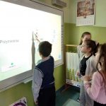 Trzy dziewczynki stoją i patrzą na tablicę interaktywną, podczas gdy chłopiec jej dotyka.