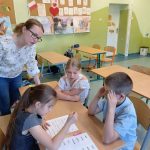 Dwie dziewczynki i chłopiec siedzą przy stoliku i patrzą na dwie kartki leżące na nim, obok nich stoi kobieta.