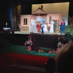 Mężczyzna w kostiumie księcia rozmawia z kobietą w białej sukni, za nimi grupa osób