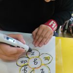 Dziewczyna przy stoliku rysuje długopisem 3d kształt kwiatka z zasadami ortografii