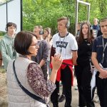 Na pierwszym planie kobieta w okularach i o ciemnych włosach, w szarej pikowanej kamizelce i wzorzystej bluzce, z uniesionymi rękami, opowiada przebieg procesów norymberskich. W tle zasłuchani uczniowie. Za uczniami drewniana konstrukcja – szubienica, na której został powieszony jeden z komendantów obozu.