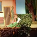 Na scenie mężczyzna w zielonym ubraniu ikoronie na głowie siedzi na podłodze