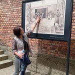 Kobieta w spodniach i szarej kamizelce, z parasolem w dłoni, pokazuje na tablicy sposób rozstrzeliwania więźniów pod tzw. „ścianą śmierci”. Tablica stoi na tle ceglanej ściany.