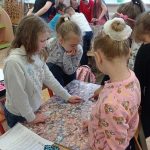 Trzy dziewczynki przyglądają się tablicy wyklejonej zdjęciami kilkoma setek główek ludzi