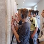 Chłopiec w szarym ubraniu i słuchawkach na uszach szuka wzrokiem znajomych nazwisk w „Księdze Zmarłych”, zajmującej przestrzeń jednego z bloków więźniarskich. Za nim inni uczniowie