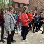 Grupa kilkunastu osób w słuchawkach na uszach stoi na chodniku i słucha opowiadania przewodniczki stoi na chodniku