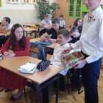 Wysoki, szczupły uśmiechnięty chłopiec w białej koszuli i ciemnych spodniach, trzyma w rękach zielono-czerwone pudełko