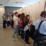 Dziewczynki i chłopcy stoją przed „Księgą Zmarłych”, symboliczną księgą ofiar obozu koncentracyjnego, zajmującą środek pomieszczenia pomalowanego na biało