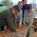 Trzech chłopców i jedna dziewczynka na tablicy wyklejonej zdjęciami głów, wskazują palcami zdjęcie głowy Miłkowskiego