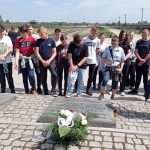 Uczniowie stoją z pochylonymi głowami przed tablicą upamiętniającą śmierć tysięcy Polaków. Przy tablicy biało-czerwone kwiaty. W tle brama prowadząca na teren obozu w Brzezince