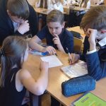 Trzech chłopców i jedna dziewczyna pochylają się nad kartką papieru. W tle kilka dziewczyn w białych bluzkach.