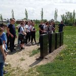 Grupa uczniów stoi przed 4 czarnymi płytami, które umieszczone są na trawiastym placu. Obok płyt kobieta w szarej kamizelce wskazuje napis na płycie.