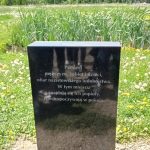Tablica z czarnego kamienia na tle jeziorka porośnietego wysoką trzciną. Na tablicy napis: „Pamięci mężczyzn, kobiet i dzieci, ofiar nazistowskiego ludobójstwa. W tym miejscu znajdują się ich popioły. Niech spoczywają w pokoju”. W tle wysokie, zielone drzewa.
