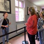 Grupa osób w sali ogląda obrazy przedstawiające tortury zadawane więźniom, przed grupą przewodniczka