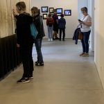 Chłopiec w czarnym ubraniu przegląda strony "Księgi zmarłych". W tle troje uczniów ogląda zdjęcia na ścianach