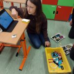 Dziewczynka z długimi włosami w okularach patrzy na tablet leżacy na stoliku. Pzed nią na podłodze żółte pudełko z drobnymi elementami. ,