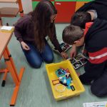 Dwóch chłopców i jedna dziewczyna pochylają się nad żółtym pudełkiem, wybierając potrzebne elementy. Dzieci siedzą na podłodze, przed nimi żółte pudełko wypełnione drobnymi klockami.