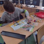 Chłopiec w szarej bluzie siedzi przy stoliku, buduje zestaw z klocków lego. Przed nim na stoliku papierowa instrukcja.