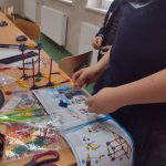 Dwóch chłopców w ciemnych podkoszulkach buduje zestaw z klocków wg instrukcji. Chłopcy stoją przy stoliku, przed nimi na stoliku leży papierowa instrukcja oraz mnóstwo drobnych kolorowych elementów. Widać też rozpoczętą budowę wciągarki.