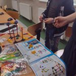 Dwóch chłopców w ciemnych podkoszulkach buduje zestaw z klocków wg instrukcji. Chłopcy stoją przy stoliku, przed nimi na stoliku leży papierowa instrukcja oraz mnóstwo drobnych kolorowych elementów. Widać też rozpoczętą budowę wciągarki.