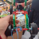 Jedna osoba trzyma w ręku pojazd zbudowany z klocków lego. dwie osoby widoczne w tle.