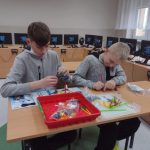 Dwóch chłopców w szarych bluzach siedzi przy stole, budując z klocków lego. Przed nimi na stoliku czwerowne pudełko wypełnione kolorowymi elementami. W tle widoczne monitory komputerów.