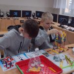 Dwóch chłopców w szarych bluzach buduje zestaw z klocków wg instrukcji. Chłopcy siedzą przy stoliku, przed nimi na stoliku leży papierowa instrukcja oraz mnóstwo drobnych kolorowych elementów oraz duże czerwone pudełko. Widać też rozpoczętą budowę wciągarki.