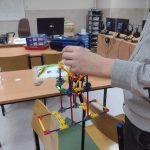 Chłopiec w szarym swetrzepokazuje zasadę działania wciągarki. w tle kolorowe pudełka z klockami do robotyki, oraz monitory komputerów.