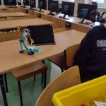 ludzik zbudowany z klocków lego stoi na stoliku na tle czarnego tabletu. W tle widać monitory komputerów oraz chłopca w ciemnym swetrze.