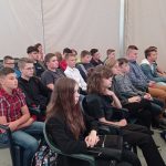 kilkunastu uczniów siedzi na plastikowych krzesłach, w tle biała ściana namiotu, w którym odbywała się konferencja