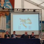 czterech mężczyzn i jeden ksiądz siedzą przy stole, jeden z nich mówi do mikrofonu. Za plecami mają ekran, na którym znajduje się mapa Europy, po lewej stronie olbrzymi baner z postacią błogosławionej Karoliny – dziewczyna ubrana jest w niebieską sukienkę, na niej ma biały fartuch. W rękach trzyma wiązkę zboża.