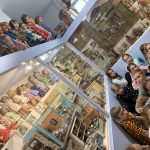 Grupa dzieci siedzi na podłodze w sali muzealnej, dookoła szklane gabloty z lalkami