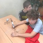 Dwóch uczniów siedzących przy ławce, jeden trzyma długopis, drugi patrzy w tył. Na ławce leży bluza.