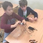 Dwóch uczniów siedzących przy ławce, jeden uczeń trzyma długopis, drugi patrzy na długopis. Przed nimi leży ładowarka.