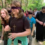 Grupa osób, na pierwszym planie dziewczyna w czarnej koszulce, ma zawiązane oczy, prowadzi ją inna dziewczyna