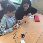 Dwie uczennice siedzące przy ławce, jedna trzyma długopis, przed nimi leżą nożyczki i piórnik.