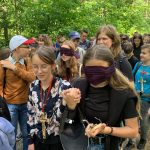 Dziewczyna z zasłoniętymi oczami prowadzona przez uśmiechniętą koleżankę, za nimi grupa osób