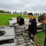 trzech chłopców przygląda się uważnie szczątkom samolotu. W tle zielona trawa.