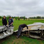 dwóch chłopców stoi przy szczątkach samolotu, trzeci chłopiec, w ciemnych spodniach i ciemnej bluzie, pochyla się nad fragmentem maszyny. W tle drzewa, krzewy i zielona trawa.
