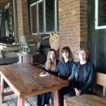 trzy dziewczynki w czarnych bluzkach siedzą przy drewnianym stole. Przed nimi talerzyk z kawałkami ciasta. W tle stare urządzenia ze wsi galicyjskiej.