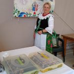 kobieta o jasnych włosach w ludowym stroju krakowskim stoi obok baneru z napisem „ Koło Gospodyń Wiejskich w Zabawie”. Przed nią pojemniki z ciastem.