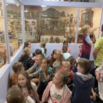 Grupa dzieci siedzi na podłodze w sali i ogląda zabawki ułożone w stojących dookoła gablotach